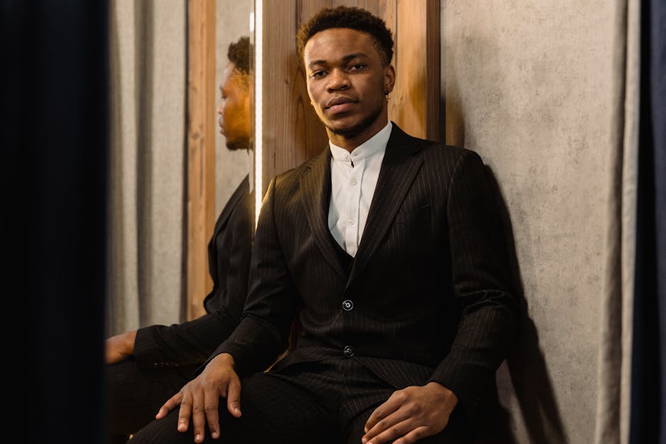 A well-dressed man in a black suit sits indoors, exuding elegance and confidence.